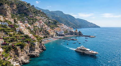 A spectacular view of the Amalfi Coast in Italy, with colorful pastel houses clinging to dramatic, steep cliffs plunging into the sparkling Mediterranean sea. The scene is captured in bright, clear daylight, emphasizing the rich blues of the water and the vibrant hues of the architecture. A luxury motor yacht cruises elegantly along the coastline. The visual style is premium, cinematic, and breathtaking, conveying a sense of exclusive European travel.