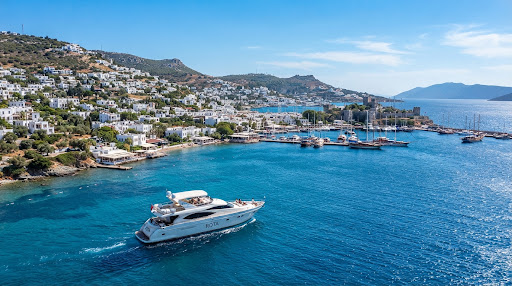 A stunning wide shot of the Bodrum coastline in Turkey on a bright sunny day. The iconic white-washed houses cascade down the hillside towards the deep blue Aegean sea. A modern, sleek luxury yacht is sailing smoothly in the foreground. The aesthetic is clean, crisp, and sophisticated, dominated by whites and vibrant blues. The atmosphere is vibrant yet elegant, capturing the essence of a premium Mediterranean summer destination.