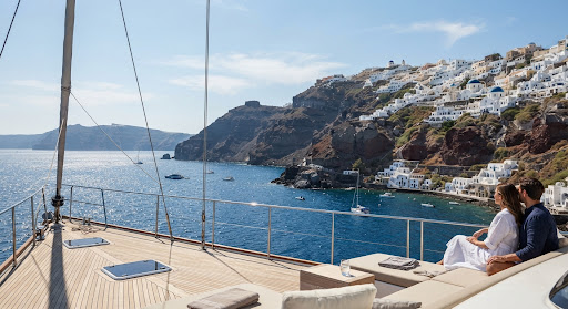 A dramatic view of the Greek islands coastline from the deck of a yacht on a sunny day. White-washed buildings cling to a steep cliffside against a backdrop of deep blue Aegean sea. The lighting is bright and clear, highlighting the contrast between the white buildings and blue sea.