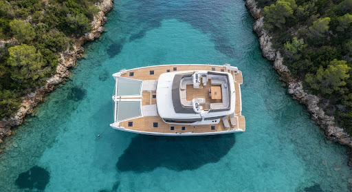 A top-down aerial view of the Azure Dream catamaran anchored in a quiet, secluded cove with turquoise water. The wooden decks are visible and spotless, with luxurious outdoor seating. The surrounding environment is lush and green, emphasizing the tranquility of a private voyage. The lighting is soft and natural, perfect for a high-end maritime brand.