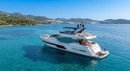 A stunning, modern luxury motor yacht, the 'Azure Horizon', anchored in the crystal-clear, sunlit turquoise waters of the Aegean Sea near Bodrum, Turkey. The yacht features sleek, aerodynamic white fiberglass lines and expansive tinted glass windows, reflecting the bright daytime sun. The background shows distant rugged, lush green coastlines under a perfect, cloudless azure sky, evoking a premium, high-end maritime lifestyle and serene luxury travel experience.