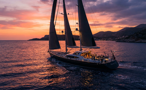 A beautiful luxury sailing yacht, the Odyssey One, gliding through the Mediterranean at sunset. Its tall masts are silhouettes against a vibrant orange and pink sky. The water is deep indigo, reflecting the twilight colors. The aesthetic is romantic yet professional, capturing the essence of high-end nautical exploration.